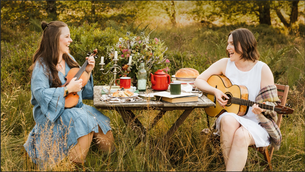 two women smiling, seated in meadow with picnic table, guitar and violin