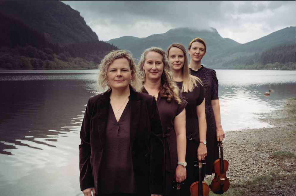 four adults standing in line holding violins on the shore of a lake with mountains in the background.